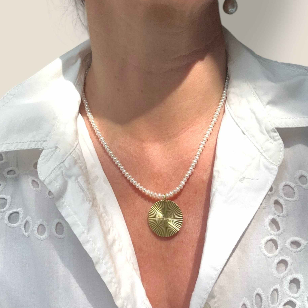 Gold Disc pendant on pearl necklace modelled against a white broderie anglaise shirt dress