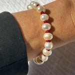 A close-up of a person's wrist wearing a beaded bracelet made of large white pearl-like beads separated by small red cord sections, with a dark sleeve visible on the left.