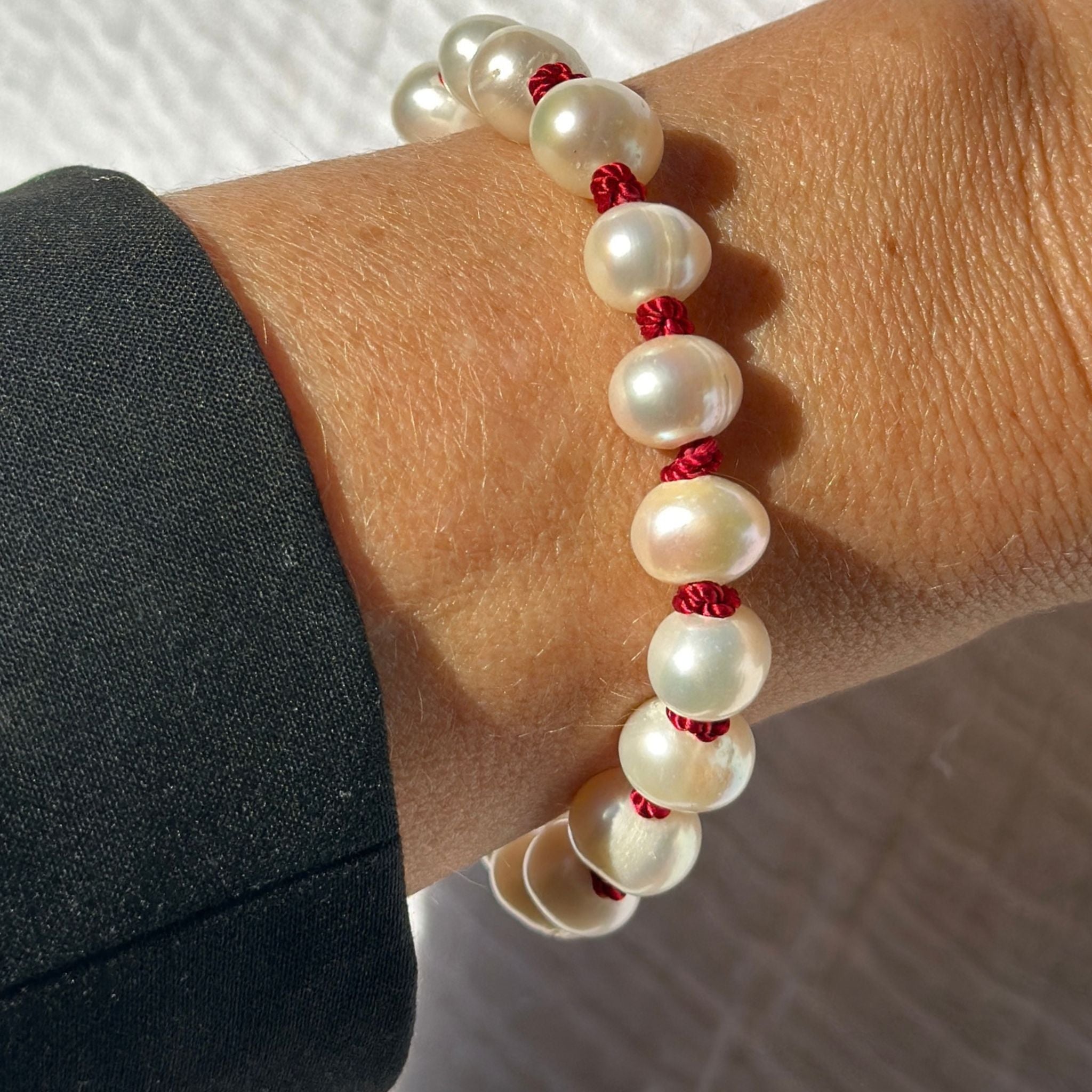 A close-up of a person's wrist wearing a beaded bracelet made of large white pearl-like beads separated by small red cord sections, with a dark sleeve visible on the left.