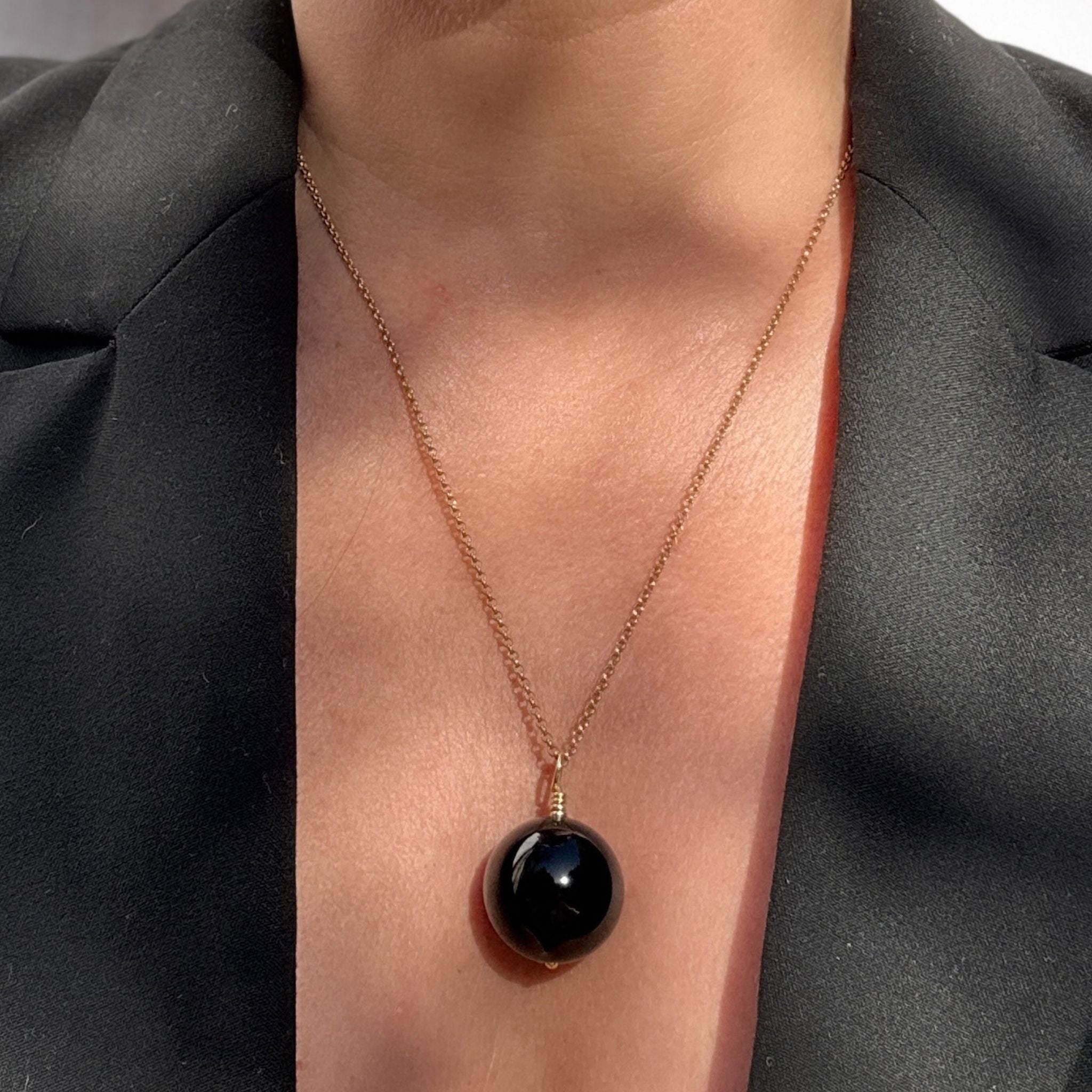 A close-up of a person wearing a delicate gold chain necklace with a large, polished black onyx pendant, styled with a black blazer.