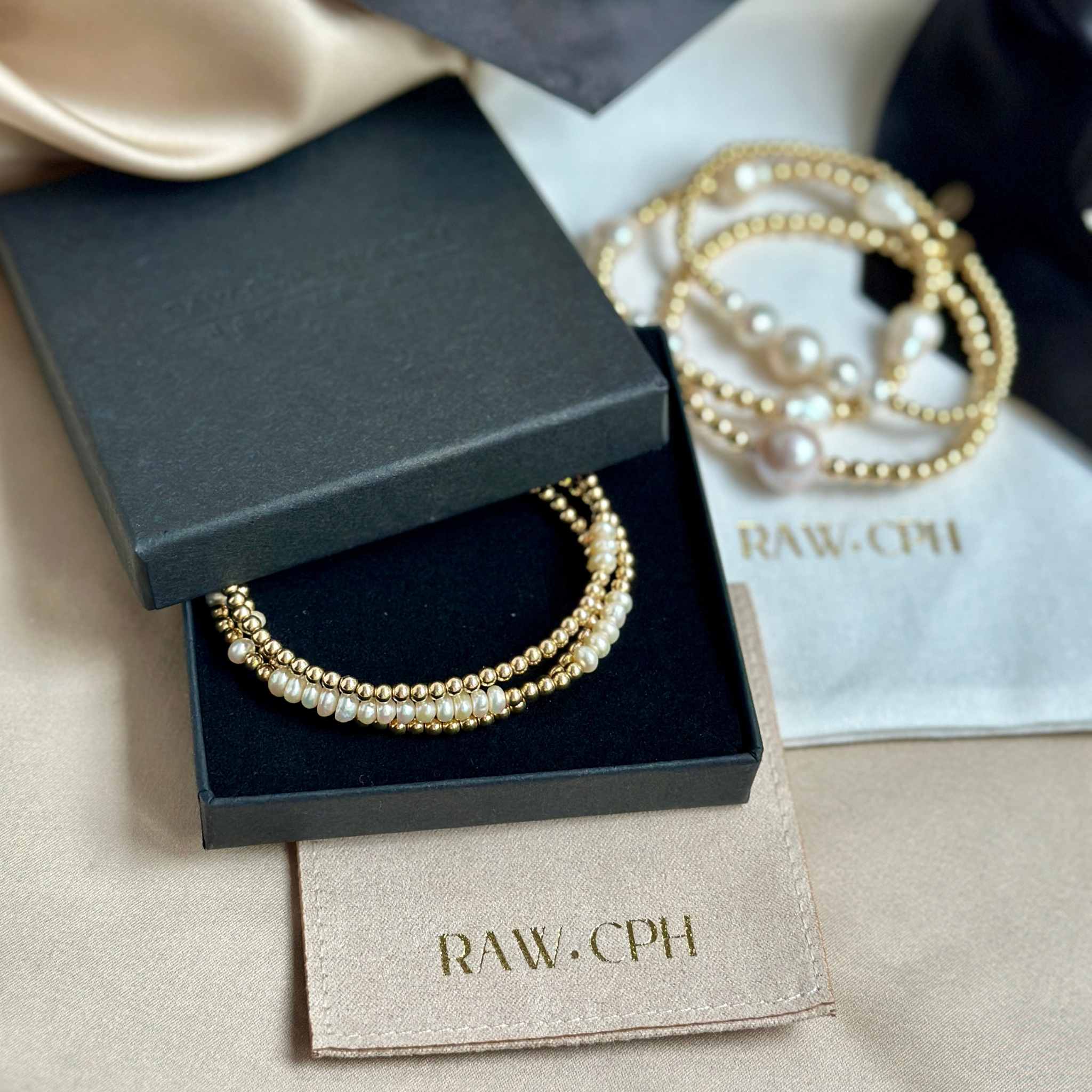 Pearl and gold bracelet in a black box with 'RAW.CPH' branding on a beige fabric background.