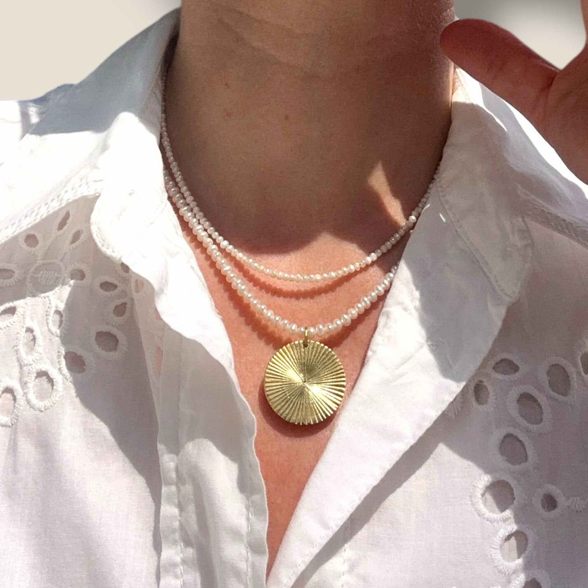 Gold Disc pendant on pearl necklace modelled against a white dress and layered with another pearl necklace
