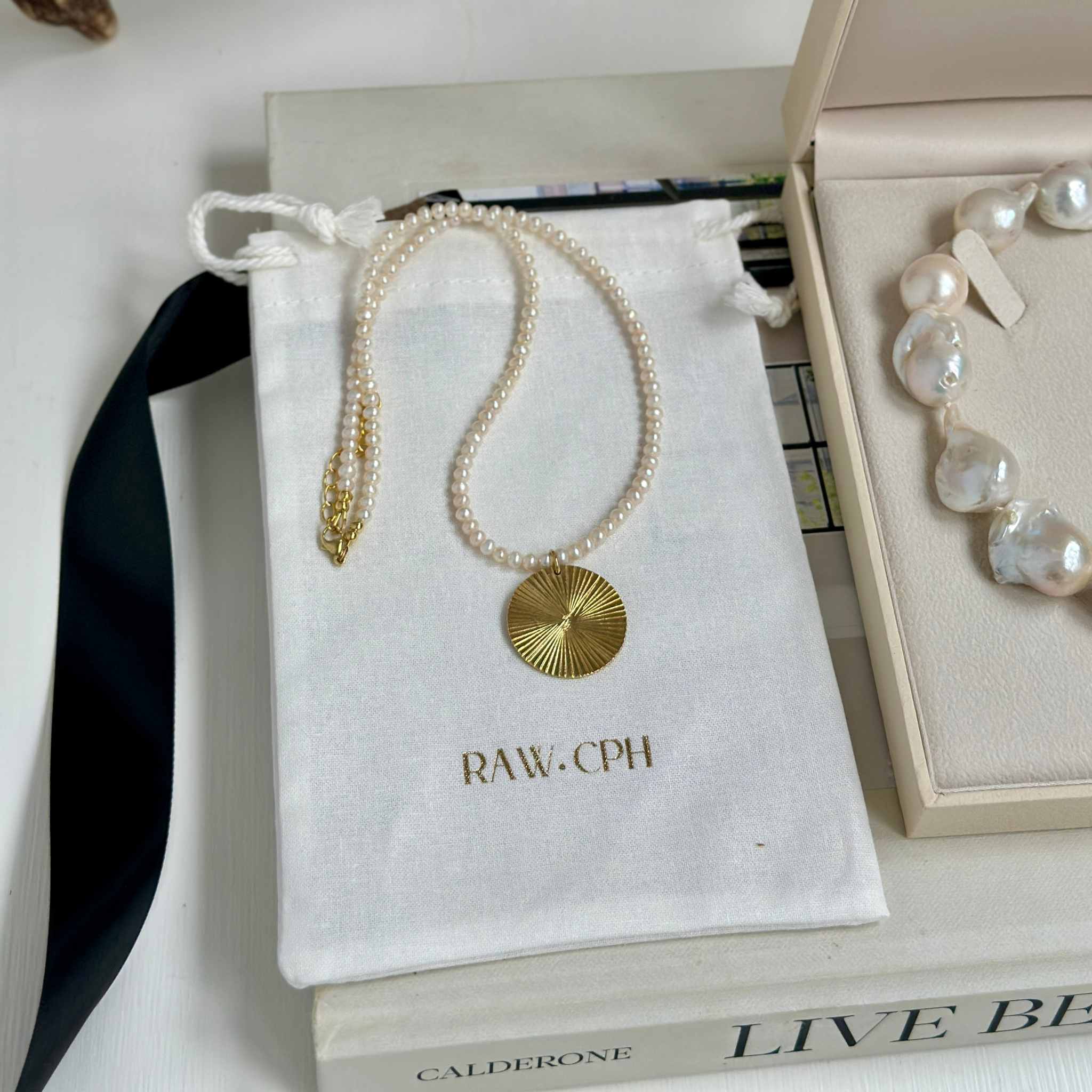 Pearl necklace with gold pendant on a branded pouch, with a jewelry box in the background.