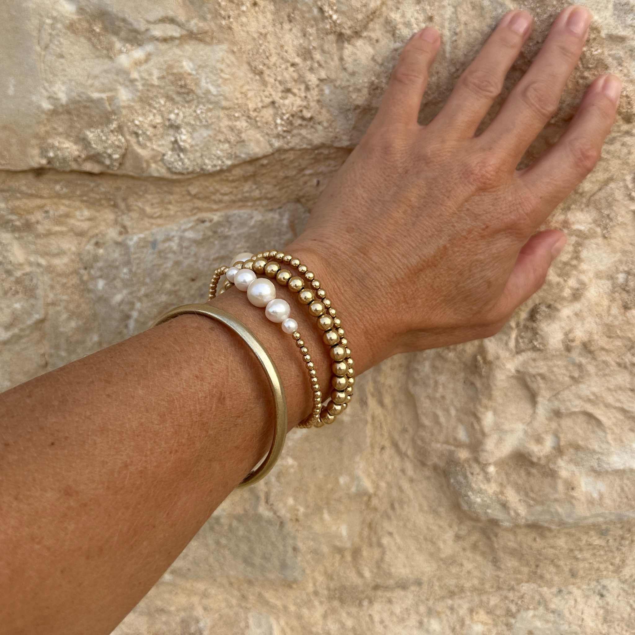 Hand wearing gold and pearl bracelets against a stone wall background