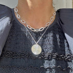 Person wearing a silver chain necklace with a circular pendant on a dark textured top.