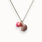 A delicate necklace featuring a gold-toned chain with a pendant consisting of two small pink beads and one larger, smooth matte pink stone bead, shown against a plain white background.