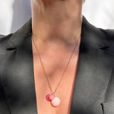 A close-up of a person wearing a delicate gold chain necklace with three small spherical charms in pink and peach tones, styled with a black blazer.