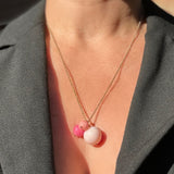 A close-up of a person wearing a delicate gold chain necklace with a pendant featuring two pink beads and one larger pale pink bead, styled with a dark gray blazer.