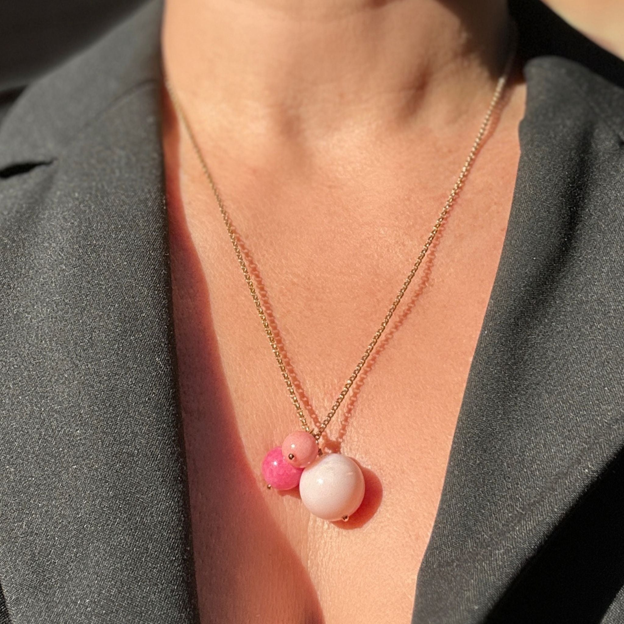 A close-up of a person wearing a delicate gold chain necklace with a pendant featuring two pink beads and one larger pale pink bead, styled with a dark gray blazer.