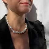 A close-up of a woman wearing a chunky baroque pearl necklace with irregular, polished white pearls, styled with a black blazer against a white textured background.