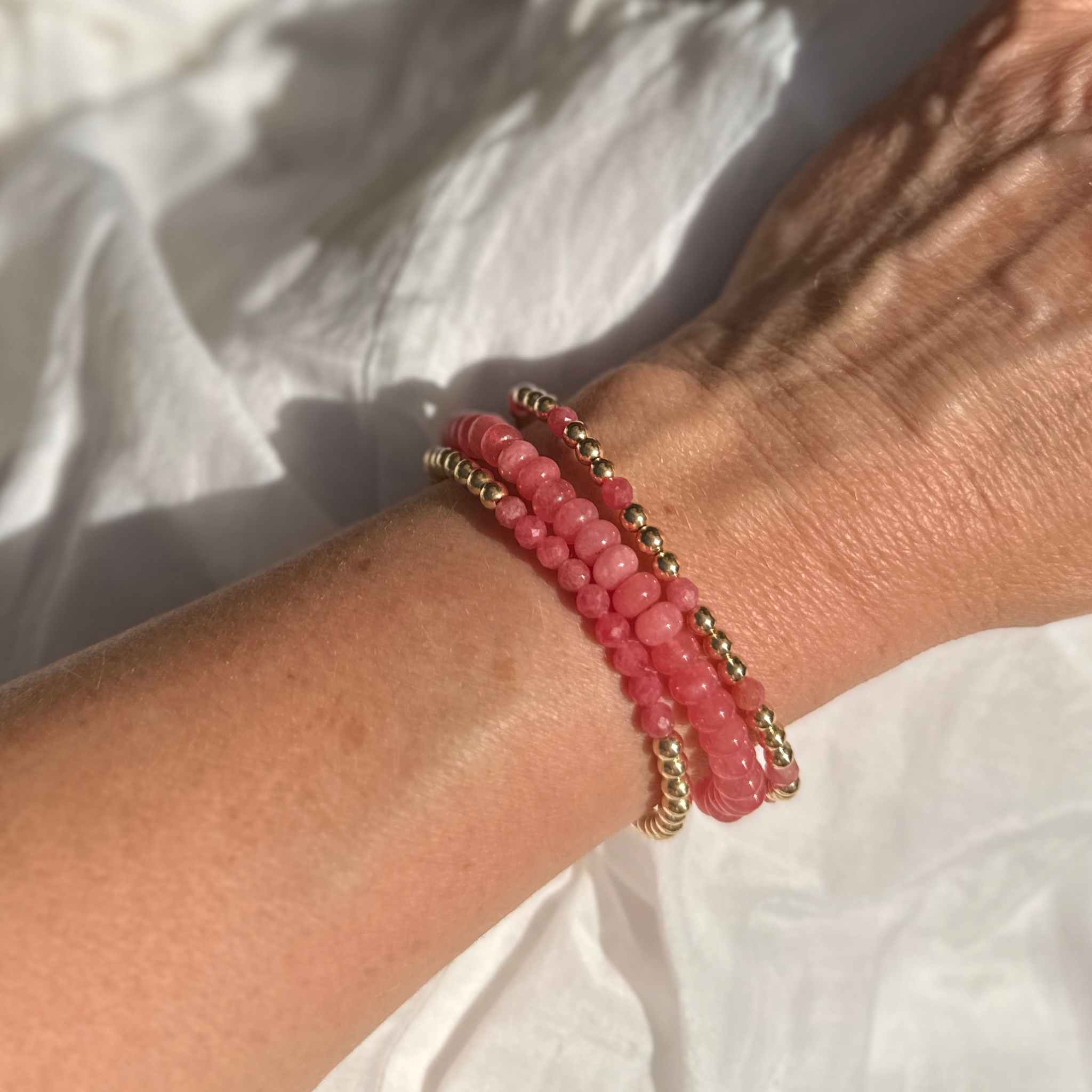 Close-up of a wrist wearing three pink rhodochrosite and jade beaded bracelets on a white fabric background