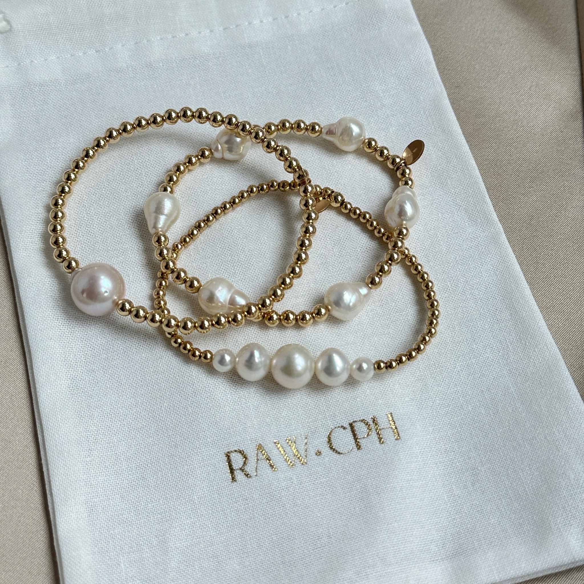 Gold bracelet with pearls on a white fabric with 'RAW.CPH' branding.